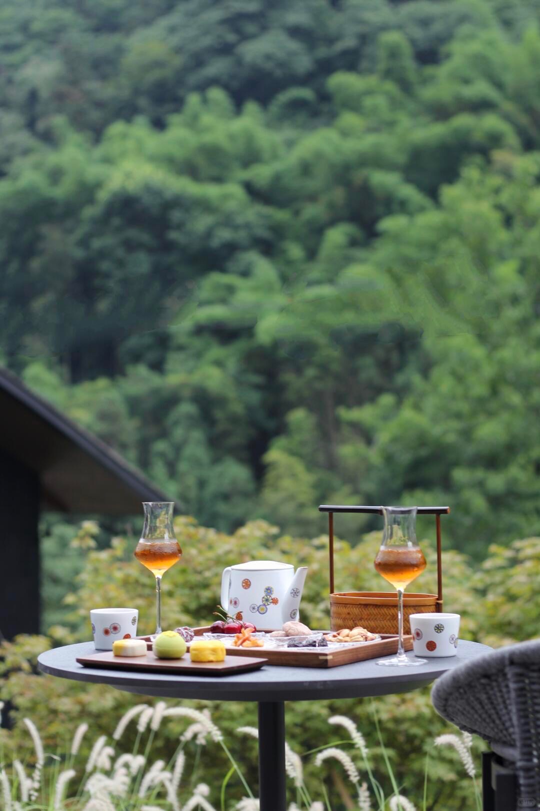 Outdoor round table with tea in stemmed glasses, teapot and a snack tray, set against a lush green forested hillside.